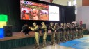 Przed sceną trwa pokaz szkoły wojskowej - mężczyźni ubrani w mundury stoją w jednym rzędzie i odgrywają scenę korzystania z broni palnej. W tle widać podświetlany napis na telebimie prezentujący nazwę szkoły.
