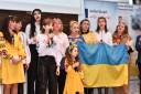 Na zdjęciu widać dziesięć osób reprezentujących ukraiński zespół, występujący na scenie. Osoby te trzymają w rękach ukraińską flagę i ubrane są w tradycyjne stroje w barwach ukraińskiej flagi. Dwie osoby stojące przed wszystkimi to dziewczynka w wieku ok 4 lub 5 lat ubrana w piękną żółtą sukienkę a na głowie ma wianek z kwiatami w kolorach ukraińskiej flagi czyli niebiesko żółte oraz dziewczynka w białej bluzce i czarnych spodniach w wieku ok 14 lat. Obie trzymają w ręku mikrofony i śpiewają
