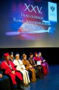 Na zdjęciu na scenie siedzą mężczyźni i kobiety ubrani w stroje i insygnia akademickie. W tle widać ekran z napisem XXV Inauguracja Roku Akademickiego 2023/2024