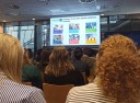 Grupa osób ogląda prezentacje na ekranie . Slajd zawiera tekst i obrazy ilustrujące różne aspekty programu Erasmus.