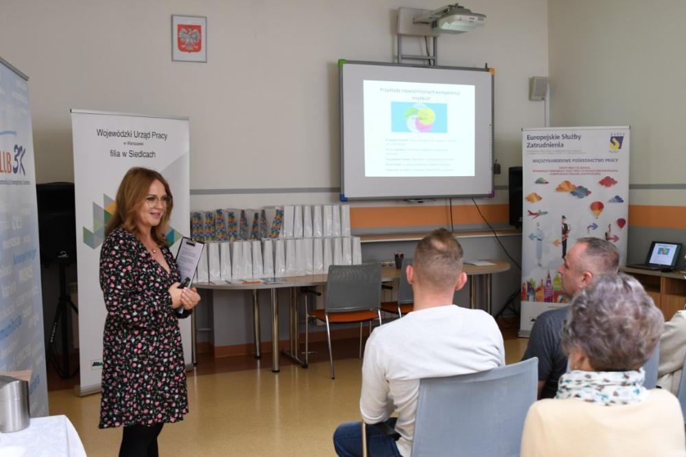 Zdjęcie galerii artykułu: Na zdjęciu znajduje się kobieta prowadząca prezentację w jasnym pomieszczeniu. Stoi ona z przodu sali, po lewej stronie, zwrócona w stronę siedzących słuchaczy. 
Za nią widać ekran projekcyjny, na którym wyświetlany jest slajd z tytułem "Przykłady najważniejszych kompetencji miękkich". Po lewej stronie ekranu znajduje się baner Wojewódzkiego Urzędu Pracy, filia w Siedlcach, a po prawej baner Europejskich Służb Zatrudnienia, dotyczący międzynarodowego pośrednictwa pracy. Na stole za prowadzącą widoczne są równo ustawione torby prezentowe.
Na pierwszym planie widoczni są uczestnicy siedzący na krzesłach, którzy słuchają prezentacji. - Pracownik Wojewódzkiego Urzędu Pracy filia w Siedlcach