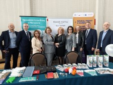 Grupa przedstawicieli WUP, PUP oraz EURES stoi przy stole wypełnionym materiałami promocyjnymi, ulotkami i gadżetami. W tle widoczne są roll‑upy informacyjne.