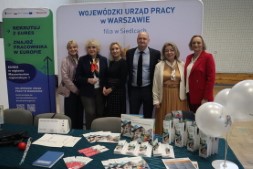 Grupa sześciu osób stoi przed ścianką informacyjną Wojewódzkiego Urzędu Pracy w Warszawie – Filia w Siedlcach. Na stole przed nimi znajdują się ulotki, broszury i materiały informacyjne ułożone w kilku stosach. W tle widoczne są roll‑upy promocyjne EURES oraz WUP.