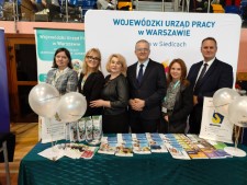 Grupa pracowników stoi za stołem z ulotkami, balonami i materiałami informacyjnymi. W tle znajduje się duża ścianka promocyjna urzędu.