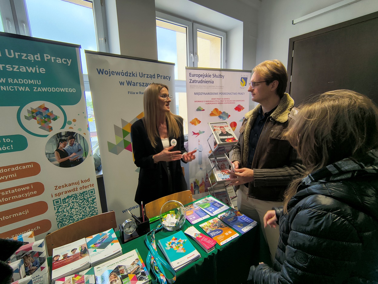 Kobieta reprezentująca urząd pracy rozmawia z młodym mężczyzną stojącym przy stoisku informacyjnym. Na stole znajdują się kolorowe broszury, notesy i materiały promocyjne. Wokół zgromadzeni są uczestnicy wydarzenia zainteresowani ofertami i poradami zawodowymi. W tle stoją banery dotyczące rynku pracy i zatrudnienia za granicą.