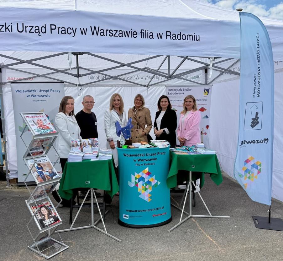 Sześć osób reprezentujących Wojewódzki Urząd Pracy w Warszawie Filia w Radomiu stoi przy stoisku informacyjnym podczas plenerowych targów pracy. Na stoisku znajdują się broszury, ulotki, czasopisma oraz małe flagi Unii Europejskiej. Nad stoiskiem widoczny jest biały namiot z napisem „Wojewódzki Urząd Pracy w Warszawie filia w Radomiu”.